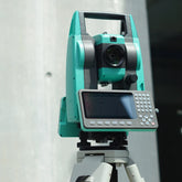 STEC total station surveying instrument mounted on a tripod, with an attached control screen and keypad, photographed against a light gray wall available to buy or hire from Tri-Site.